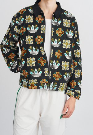 Mand iført sort lynlåstrøje med farverige geometriske og blomsterprint, kombineret med hvide bukser med et grønt logo.