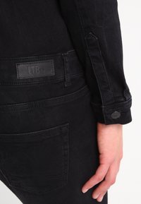 Černé džíny z denimu s hladkou texturou, opatřené malou nášivkou s logem a detaily s knoflíky na manžetě, s pohledem na zadní kapsu.