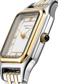 L'orologio rettangolare Rosefield presenta una cassa in metallo argento e oro, un quadrante bianco con dettagli dorati e una corona nera. Il cinturino è composto da una combinazione di bracciali in oro e argento.