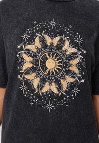 Black t-shirt featuring a circular graphic with butterflies and a sun at the center. Gold and white text and star accents throughout.