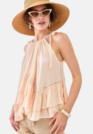 Joven mujer con top beige con volantes y cuello halter, sombrero de ala ancha, gafas de sol estilo ojo de gato, pantalones beige y pulsera dorada, posando contra fondo blanco.