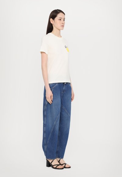 Cremfarbener T-Shirt mit kurzen Ärmeln und gelbem Grafikdesign, kombiniert mit lockeren blauen Jeans aus Denim mit schrägen Nähten und schwarzen Absatzsandalen.
