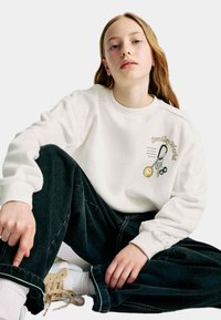 Sweatshirt blanc avec un graphique "Smiley World" et un détail sourire jaune ; porté avec un pantalon en denim bleu foncé et des baskets claires. Coupe décontractée.