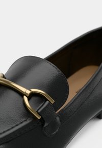 Mocassin en cuir noir avec bout arrondi et détail en métal doré en forme de mors de cheval à l'avant, présentant une texture lisse et un talon bas.