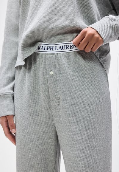 Άτομο που φοράει ανοιχτό γκρι παντελόνι και μπλούζα από ύφασμα με υφή βάφλας, σηκώνει το πουκάμισό του για να δείξει τη λευκή ζώνη μέσης με τη λέξη «RALPH LAUREN» σε σκούρα γράμματα.