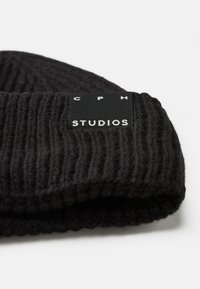 Melnā adīta cepure ar ribotu tekstūru, kurai ir taisnstūrveida birka ar baltiem uzrakstiem "CPH" un "STUDIOS". Mīksts vilnas materiāls.