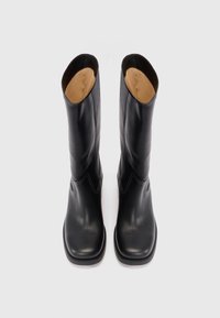 Las botas de cuero negro hasta la rodilla cuentan con una textura suave, punta redondeada, costuras en contraste y una curva sutil en el borde superior.