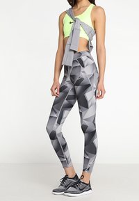 Neongrön sport-BH, grå hoodie knuten i midjan, och tajta leggings med ett geometriskt grått mönster. Svarta löparskor fulländar looken.