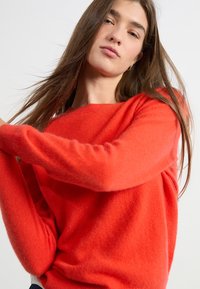 Pull en tricot rouge à manches longues, avec un col large et une texture douce. Le pull tombe librement autour du corps.