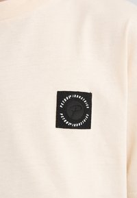 Lichtbeige katoenen T-shirt met een zwart vierkant patch waarop de tekst "PETROL INDUSTRIES" staat en een cirkelvormig logo in het midden.