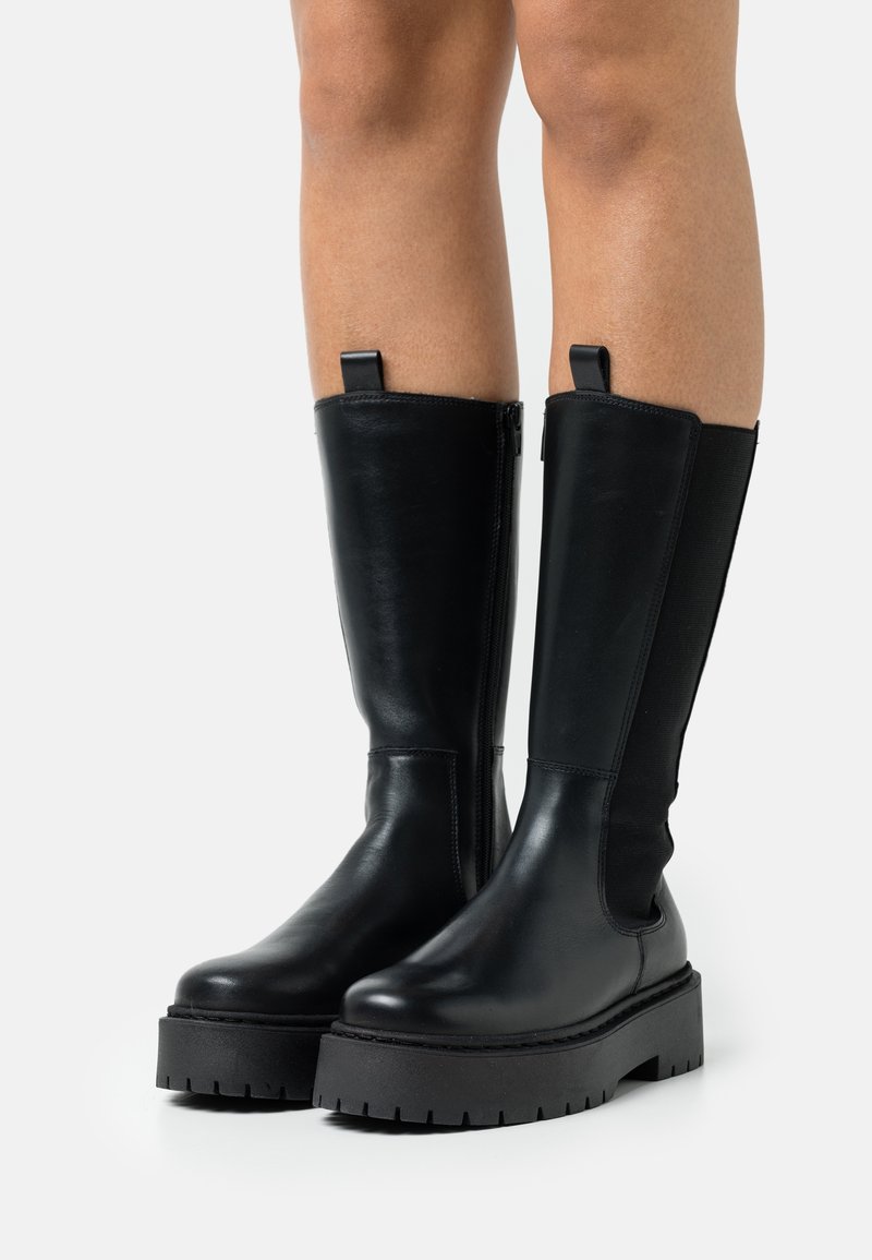 Bottes en cuir noires montantes jusqu'aux genoux avec semelles épaisses à crampons portées par une personne, avec une fermeture éclair latérale et une languette de tirage en haut, sur fond blanc.