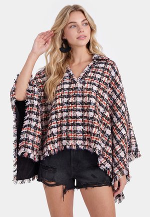 Giovane donna che indossa un poncho a quadri multicolore con frange e pantaloncini di jeans neri strappati, posando con la mano vicino alla testa.
