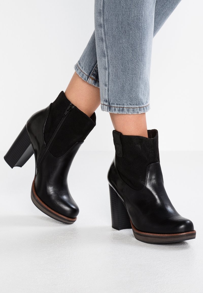 Bottines noires à la cheville avec une finition en cuir brillant et un dessus en daim. Elles sont dotées d'un talon épais et d'une fermeture éclair latérale pour un enfilage facile, portées avec un jean court.