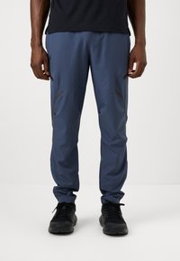 Pantalons de sport pour hommes en tissu bleu foncé. Présentent un design fuselé, des accents gris, des poches latérales et une taille élastique. Associés à des baskets noires.
