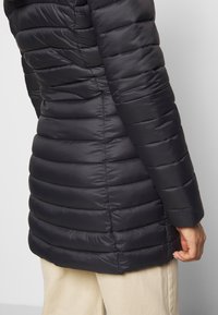 Schwarze Pufferjacke mit horizontalen Steppnähten, die eine glatte Textur und eine taillierte Passform aufweist. Die rechte Hand liegt auf dem Saum der Jacke.