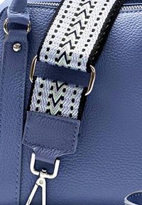 Detail van een blauwe leren tas met een gevlochten schouderriem met een zigzagpatroon in zwart en wit, zilverkleurige hardware en een gestructureerde afwerking.