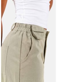 Beige cargobroek met een elastische tailleband, zijzakken en detail van klepzakken. De stof lijkt stevig en heeft een gladde textuur.