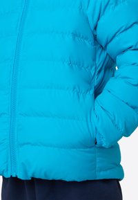 Μπλε μπουφάν με καπιτονέ υφή, κλείσιμο με φερμουάρ και πλαϊνές τσέπες. Εξοπλισμένο με ψηλό γιακά και ελαστικά μανίκια για επιπλέον ζεστασιά.