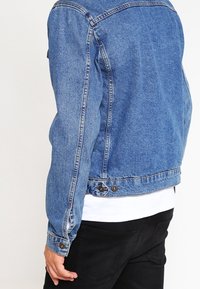 Sininen farkkutakki istuvaan muotoon. Varustettu napitettuilla hihansuilla, sivusaumoilla ja pyöristetyllä helmalla. Päällä valkoinen paita.