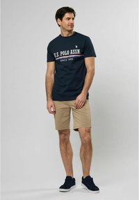 Tamno plava pamučna majica s bijelim i crvenim tekstom U.S. Polo Assn. Uparen s bež šortsevima i tamnoplavim tenisicama, stoji naslonjen na svijetlu pozadinu.