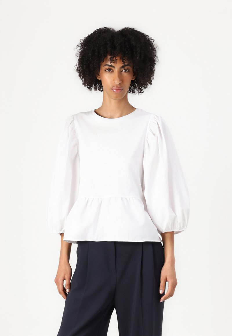 White peplum top with puffed sleeves, round neckline, and smooth texture. Paired with dark, tailored trousers.