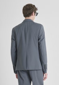 Sivi sako, prilagođenog kroja, glatke teksture, s jednim prorezom straga, detaljem s četiri gumba na rukavima, nosi se s naočalama.