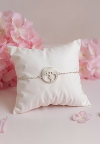 Silverfärgad armband med en rund charm som har två babysteg. Visas på en mjuk, vit satinpillow mot en ljus bakgrund.