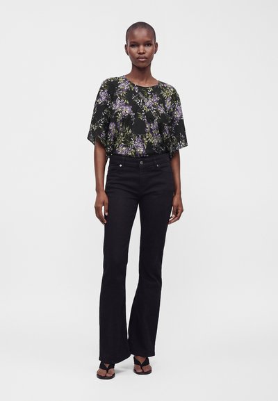Femme debout portant un chemisier noir à fleurs violettes, un pantalon flare noir taille haute et des sandales noires à bout ouvert sur fond blanc.