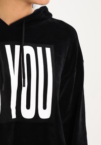 Schwarzer Kapuzenpullover aus weichem Material mit einem großen weißen Grafik "YOU" auf der Vorderseite, mit verstellbaren Kordeln am Hals.