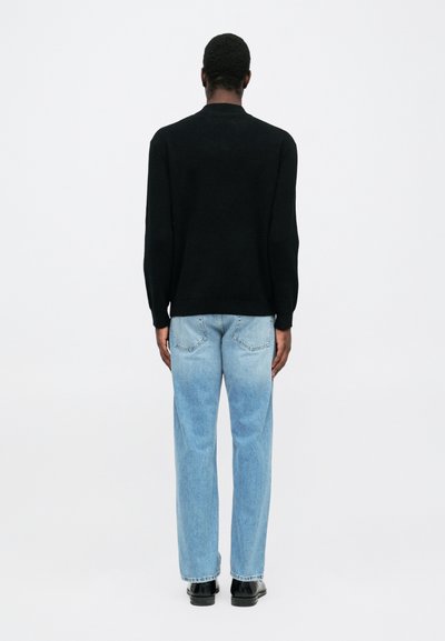 Uomo di spalle che indossa una maglietta nera a maniche lunghe, jeans blu chiaro e scarpe nere su sfondo bianco semplice.