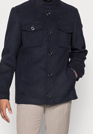 Veste boutonnée bleu marine avec deux poches à rabat à l'avant, une main dans la poche latérale, portée avec un pantalon beige clair.