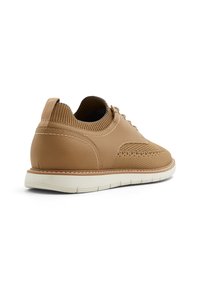 Zapatilla informal de piel color marrón con cuello de malla tejida, diseño de cordones, detalles perforados y suela de goma blanca. La vista trasera muestra un acabado texturizado.