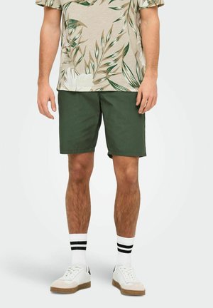 Grønne shorts med flad front i let stof. Kombineret med en beige blomster t-shirt og hvide sneakers med sorte striber på sokkerne.