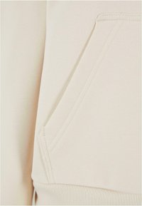 Détail d'un sweatshirt beige présentant une poche kangourou à l'avant, un ourlet côtelé, et une texture de tissu lisse avec des coutures visibles.