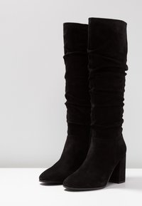 Svarta mockaskor i knähöjd med en avslappnad design, spetsig tå och en kraftig klack. Slät yta med minimal synlig hårdvara.