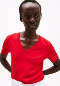 Raudonas medvilninis V formos kaklo marškinėliai, arti kūno prigludimo, su mažo logotipo akcentu. Lygus paviršius ir trumpomis rankovėmis.