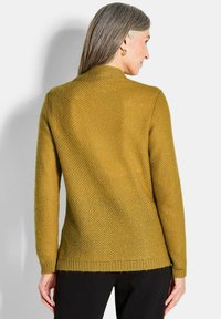Strickpullover in Senfgelb, mit einem strukturierten Muster, langen Ärmeln, Rundhalskragen und einem lässigen Schnitt, kombiniert mit schwarzen Hosen.
