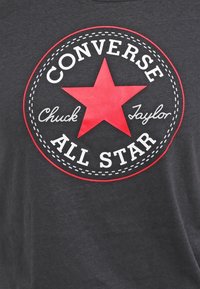 Camiseta negra con un gran gráfico circular que incluye una estrella roja, el texto "CONVERSE", "Chuck Taylor" y "ALL STAR" en blanco.
