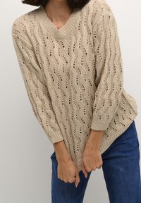 Pull beige en maille avec col en V et coupe ample, présentant un motif ondulé et des poignets côtelés. Porté avec un jean en denim bleu.