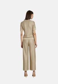 Femme debout de dos, portant un haut court beige texturé assorti et un pantalon large beige avec des sandales à talons hauts beiges.