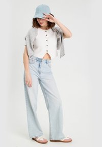 Crop top blanc avec des boutons en argent, cardigan gris drapé sur les épaules, jeans larges bleu clair, et baskets beiges avec des accents rose.
