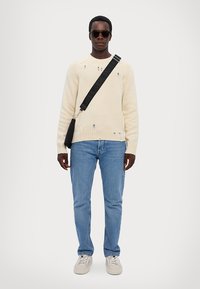 Pulover crem tricotat cu detalii distruse, blugi albaștri deschiși, ochelari de soare negri și o geantă crossbody neagră, purtat cu adidași bej.