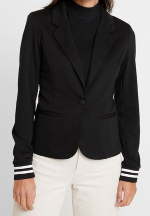 Femme portant un blazer noir ajusté avec fermeture à boutons et poignets rayés par-dessus un col roulé noir côtelé et un pantalon blanc.