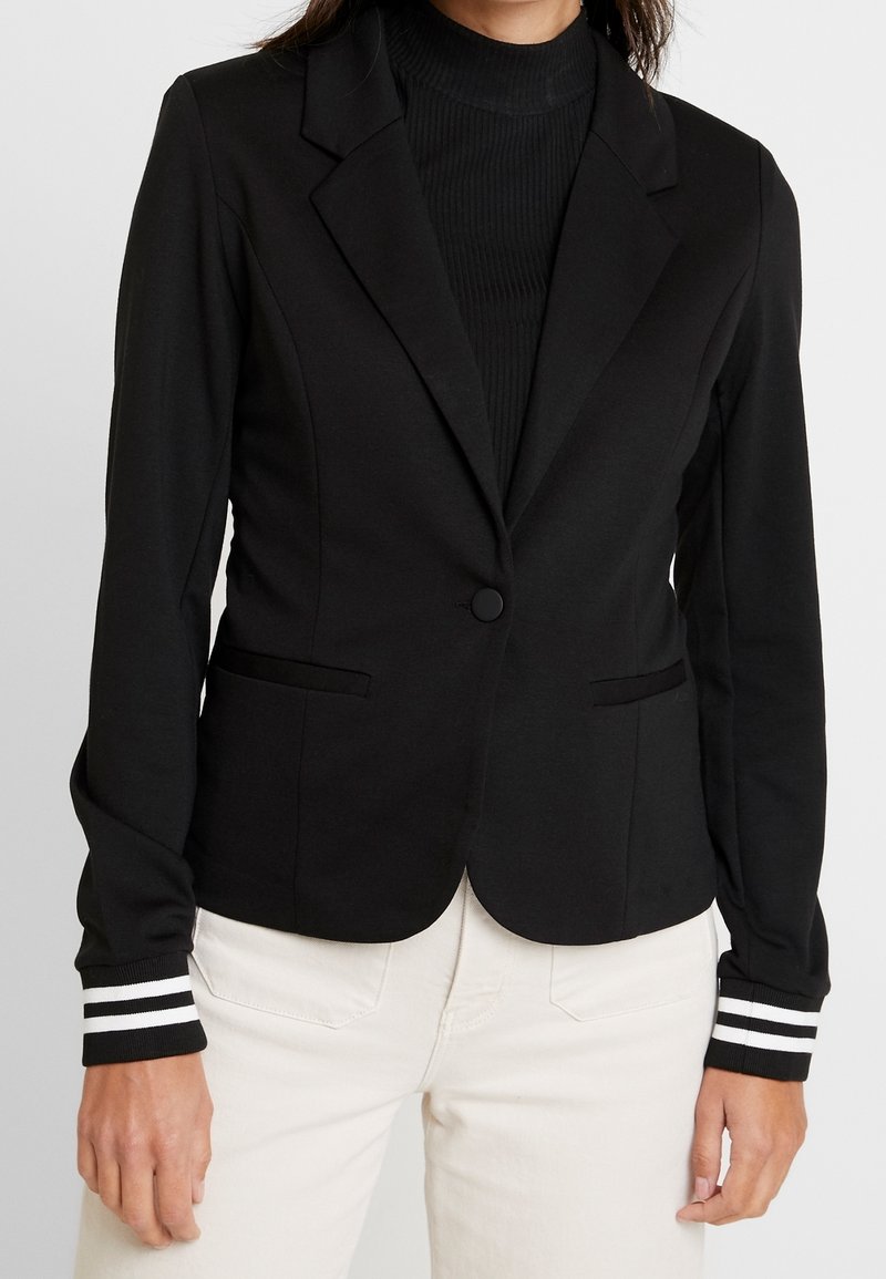 Femme portant un blazer noir ajusté avec fermeture à boutons et poignets rayés par-dessus un col roulé noir côtelé et un pantalon blanc.