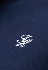 Υφασμα σε ναυτικό μπλε με λευκά κεντημένα διασταυρωμένα αρχικά "T" και "H" σε καλλιγραφικό στυλ.