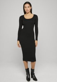 Robe en maille c ôtelée noire avec un col rond, des manches longues et une silhouette moulante, atteignant le milieu du mollet. Portée avec des bottines noires.