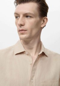 Beige linen shirt with a button-down collar, single chest pocket, and smooth texture. The shirt is worn by a male model against a plain background.