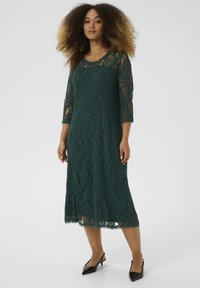 Lace midi dress in dark green, featuring three-quarter sleeves, scalloped hem, and intricate floral patterns. Styled with black heels.