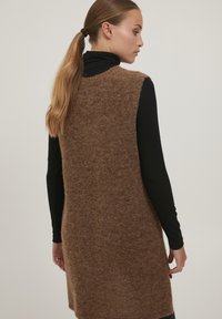 Brun strikket ærmeløs sweater med blød struktur, løs pasform og buet kant; bæres over en tætsiddende sort ribbenet turtleneck.