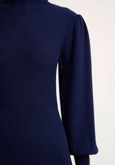 Marineblauwe geribbelde tricot jurk met een hoge kraag, gepofte lange mouwen en een getailleerd lijfje dat overgaat in een uitlopende rok.